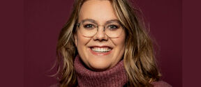 Smiling woman with glasses and wavy blond hair in a burgundy polo-neck jumper, against a matching plain background