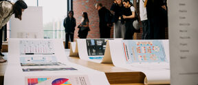 People in a hall viewing large infographic posters spread across wooden steps by a brick wall.