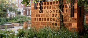 Perforated orange brick wall structure in a lush city garden with tall grass and trees