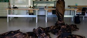 A person wrapped in brown fabric stands in a studio while the fabric spreads in folds across the floor.