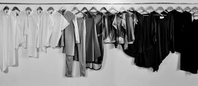 Row of minimalist shirts, tunics and trousers on hangers, fading from light to dark tones against a plain wall