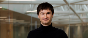 Smiling man in a black turtleneck stands indoors before glass walls and bright ceiling beams