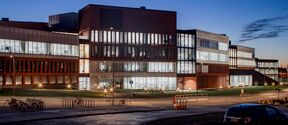 Modern glass and brick office building lit up at dusk, with parked cars and bicycles in front