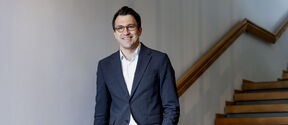 Smiling man in a dark suit and glasses stands by a wooden staircase in a bright hallway