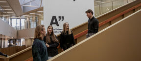 Four students stand on an indoor staircase, talking under a large Business Better Society sign.