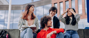 Four students sitting on outdoor steps, talking and laughing with books and a tablet by modern glass buildings