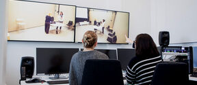 Two people at computers monitor a lab scene on large wall screens, one pointing at the video.
