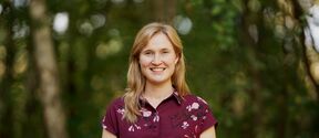 Smiling woman in a patterned burgundy shirt standing outdoors against blurred trees