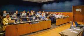 Small audience in a modern lecture hall, seated with laptops and water bottles, facing a table at the front