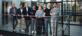 Nine people stand on an indoor metal balcony in smart casual clothes, posing for a group photo.