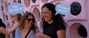 Two laughing women chat in speech bubbles outside a pink wall with round openings, text unite!
