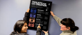 Two smiling women hanging a framed poster on the wall. The poster is an info poster of the Toolkits for Co-Creation.