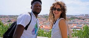Två leende turister med ryggsäckar vid en utsiktsplats, stadens tak och grönska i bakgrunden