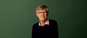 Middle‑aged man with glasses seated, wearing dark jumper and pink shirt, against plain green background.