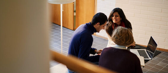 Kolme ihmistä istuu lattialla käytävässä kannettavan kanssa keskustellen.