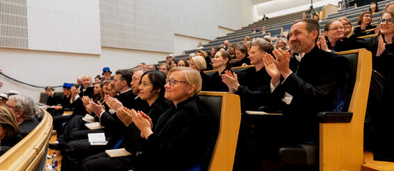 An audience wearing formal attire clapping in a modern lecture hall with tiered seating and wooden accents.