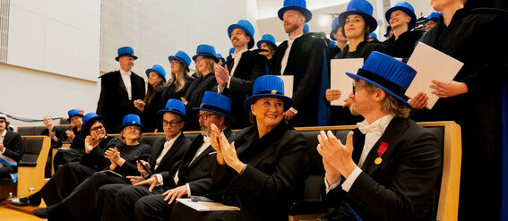 Group of people at a graduation ceremony wearing black academic robes and blue hats, some seated and some standing, clapping.