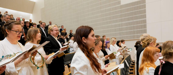 People in white and holding papers are seated and standing in rows in a modern auditorium.