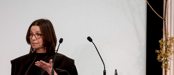 Person in dark clothing standing at a podium with two microphones, giving a presentation.