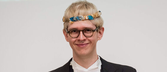 Person wearing formal black suit with white bow tie and holding a certificate. Blue hats can be seen in the background.