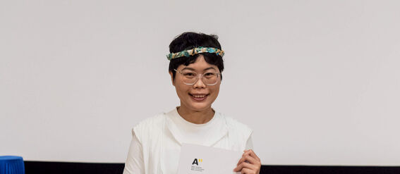 Person in white attire holding a document in an academic ceremony, with blue academic hats on the table.