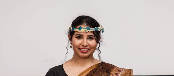 Person in a brown sari holds a white certificate in front of a table with blue academic caps.