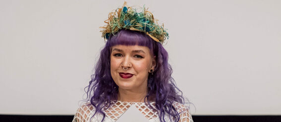 A person with purple hair wearing a white net-like dress and holding a document. Blue hats are visible in the background.