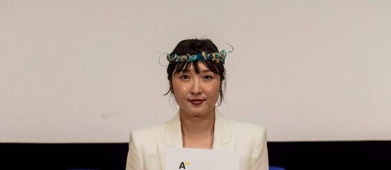 A person in a white suit holds a certificate. Blue caps are on a table in the background.