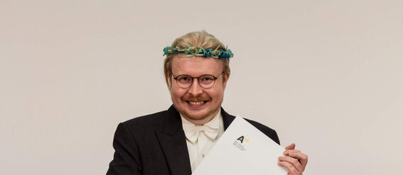 Person in formal attire holding a diploma, with multiple blue academic hats on the table behind.