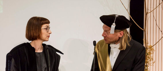 Two people dressed formally stand at a podium speaking into microphones. One wears a sash and cap.