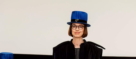 Person in black academic regalia holding a certificate with blue hats in the background.