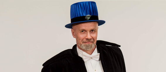 A person in academic dress with a blue hat and black robe holding a document. Several blue hats are on a table behind.