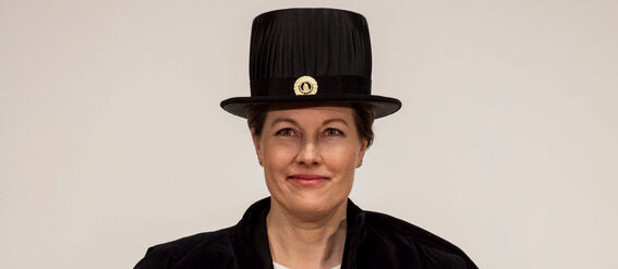 Person in academic robe and hat holding a diploma. Another hat in the background.