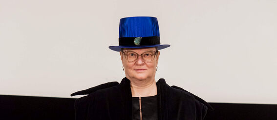 Person wearing a black academic gown and a blue academic hat, holding a certificate with more blue hats in the background.