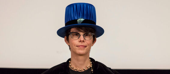 Person wearing ceremonial attire with a blue hat, holding a diploma in front of a screen