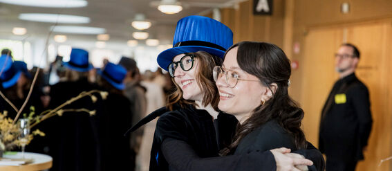 Two women hugging at an academic ceremony, one wearing a blue hat and cape.