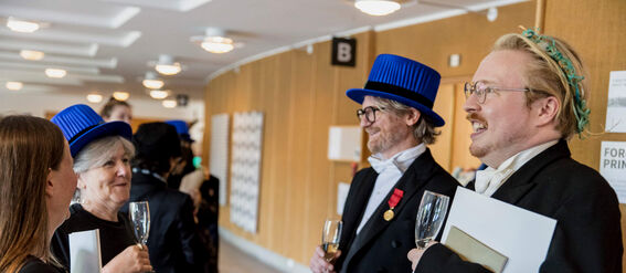 Several people standing indoors, some wearing academic regalia with blue hats, holding glasses and documents.