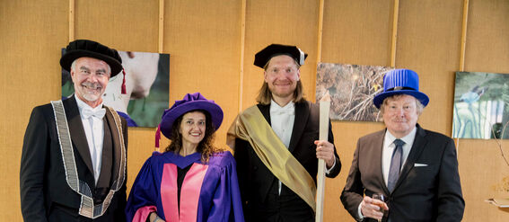 Four people dressed in ceremonial academic attire, standing indoors with artwork on the walls.