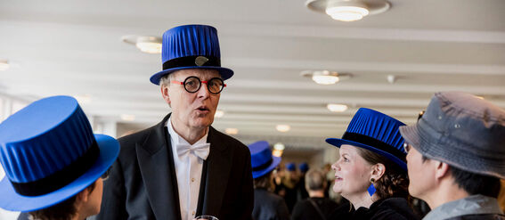 Group of people in formal attire with blue academic hats, socialising and holding drinks in a well-lit room.