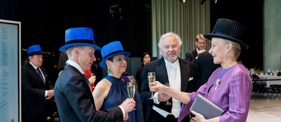 Four people toast with champagne at a formal event. Two wear bright blue hats, one wears a black hat, and one is in formal attire.