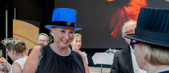 Group of people at an event, wearing formal attire and hats. A woman in a black dress holds a champagne glass.