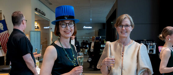 Two women in formal attire holding champagne glasses at an event. One wears a blue hat, the other a beige jacket.