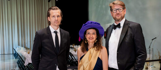 Three people dressed formally with one wearing a unique blue hat and gold sash. A decorated banquet table is in the background.