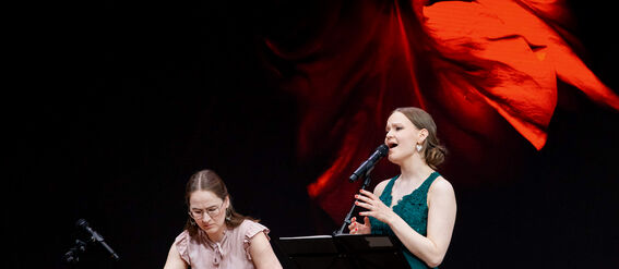 Two women performing: one plays a wooden string instrument while another sings under a backdrop of red abstract art.