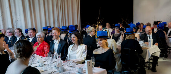 Formal banquet with guests wearing blue hats seated at white tablecloth-covered tables.