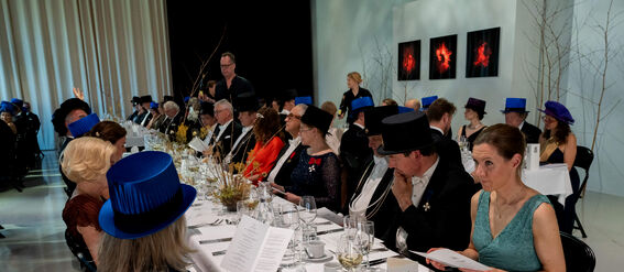 People in formal attire, including blue caps, seated at a long banquet table with menus and decorations.