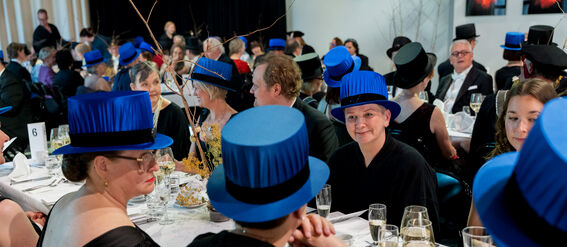A formal event with people wearing black attire and blue academic hats sitting at a white-clothed table.