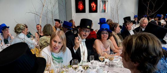 A formal gathering with people in suits and dresses, wearing black and blue hats, seated around a table with drinks.