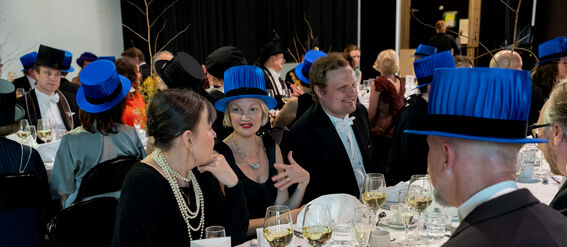 Formal gathering with people wearing blue academic hats and tuxedos, seated at tables with wine glasses.