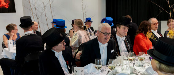 A formal banquet with several people wearing black tailcoats and ceremonial hats, seated around tables with white tablecloths.